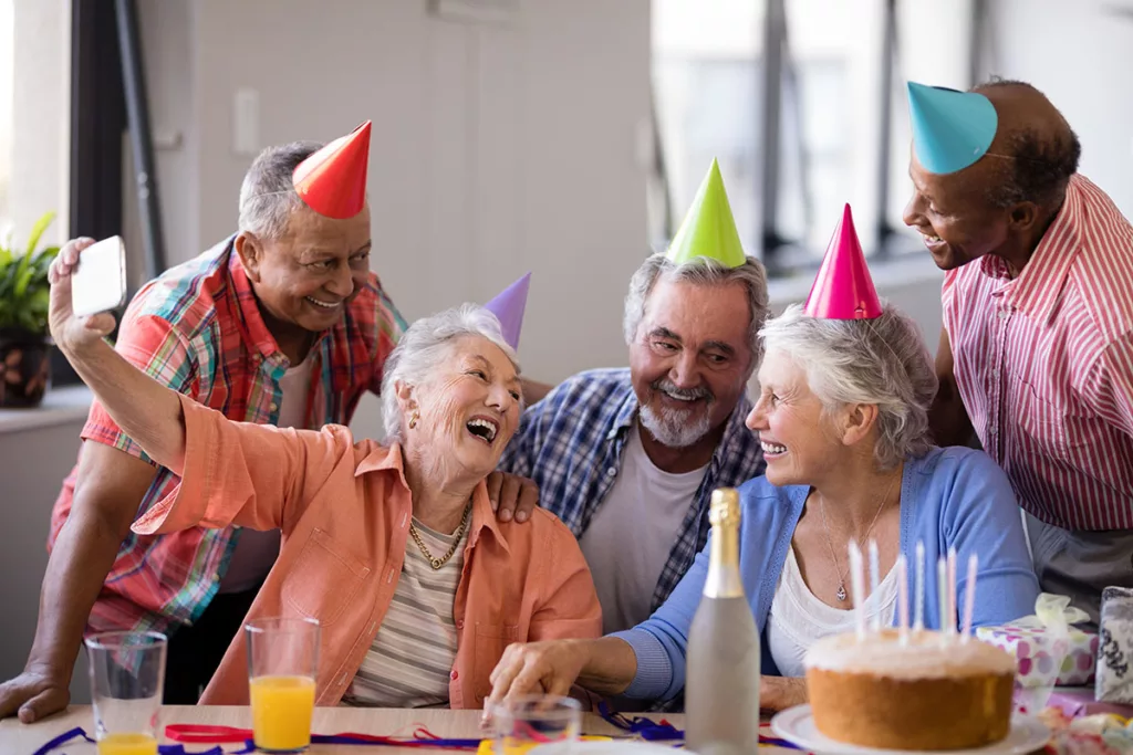 Seniors at birthday party wearing birthday hats and smiles as woman in group takes selfie. 