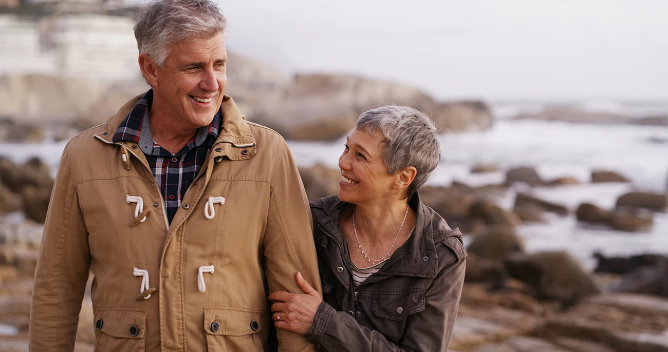 Happy older, married couple walking along a rocky, seaside beach laughing and holding hands